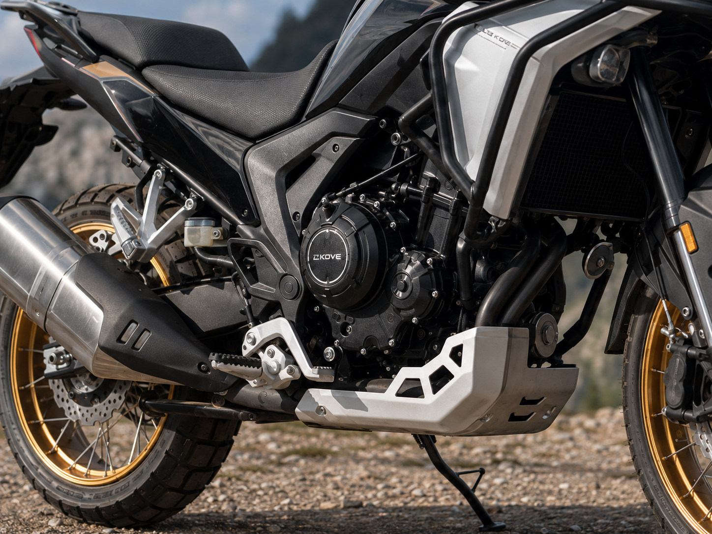 Primer plano del motor, escape y protector cárter de la Kove 625X con paisaje de montaña de fondo